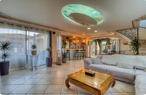 A spacious living room with a large sofa, a wooden coffee table, and a ceiling with a unique design. There is a bar area in the background and a staircase leading to another level. - Villa Le Rossignol - Vacation Home for 10 People ⭐️⭐️⭐️⭐️⭐️ (Puimisson)
