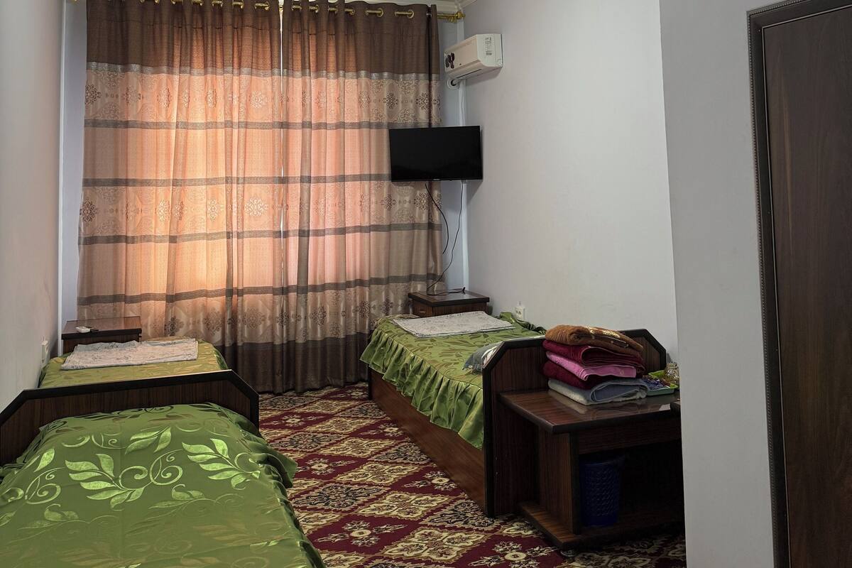 Una stanza piccola e luminosa con un letto singolo, un comodino, una televisione e una finestra con tende.