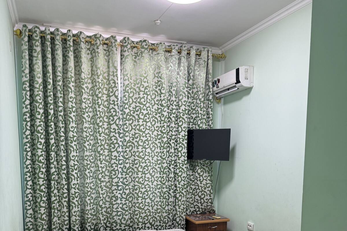 Una stanza con un letto, un comodino in legno, una TV a parete e una tenda decorata.