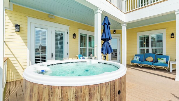Un bain à remous en bois sur une terrasse en bois, avec un parasol bleu, une véranda blanche avec un canapé bleu et un bâtiment jaune aux contours blancs.