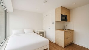 A bedroom with a bed, a small desk, a door, and a kitchenette with a microwave and sink.