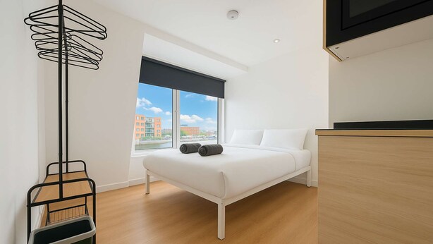 A modern bedroom with a bed, a wooden floor, a window with a view of buildings, and a black metal floor lamp.