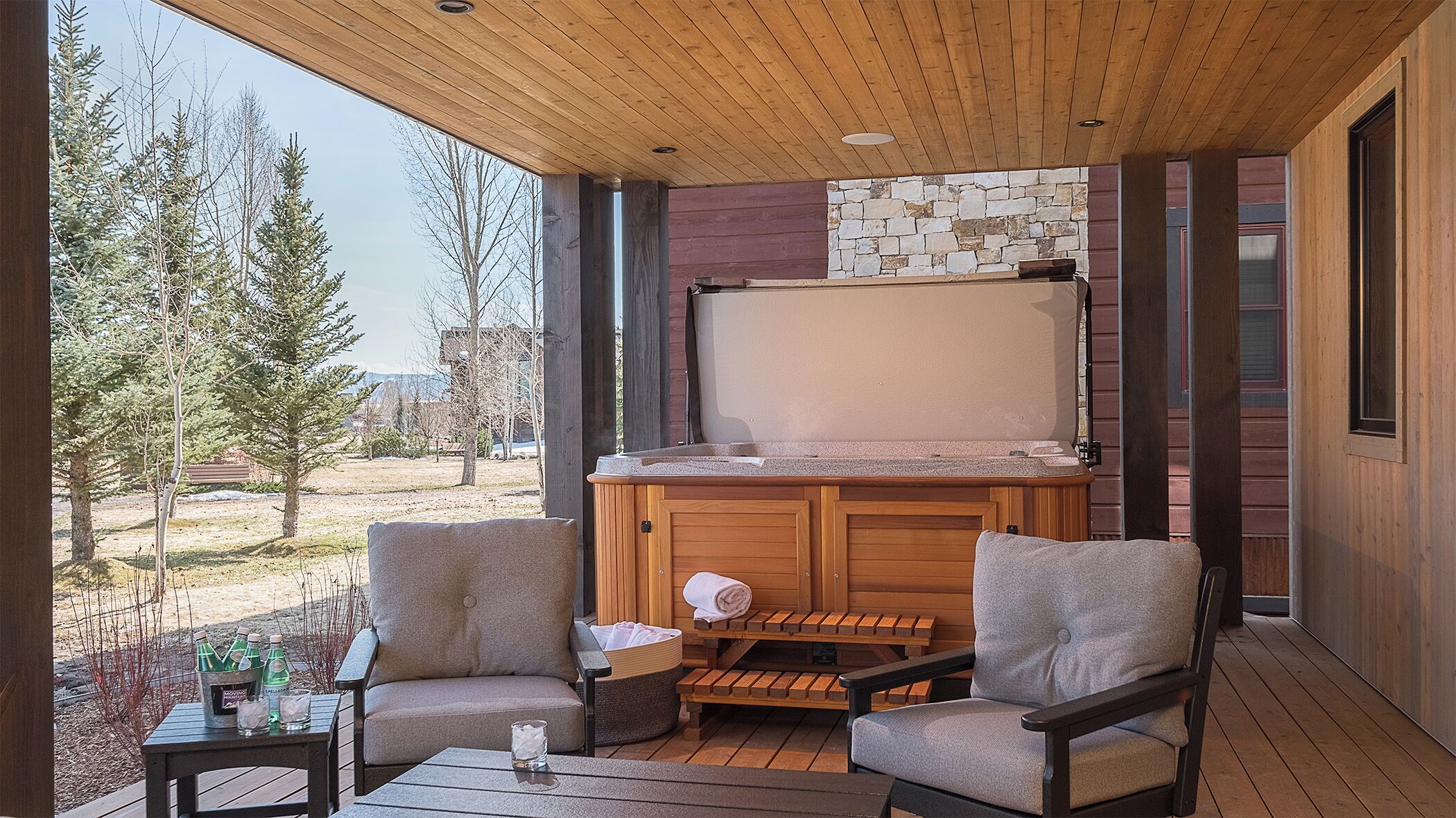 A covered outdoor area with a stone fireplace, wooden furniture, and a view of trees and buildings.