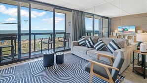 A modern living room with a large window overlooking the beach, a sofa with striped cushions, and a television on the wall. - Family Friendly condo w/ Pools by Seaside Vacations & Sales (Ocean City)