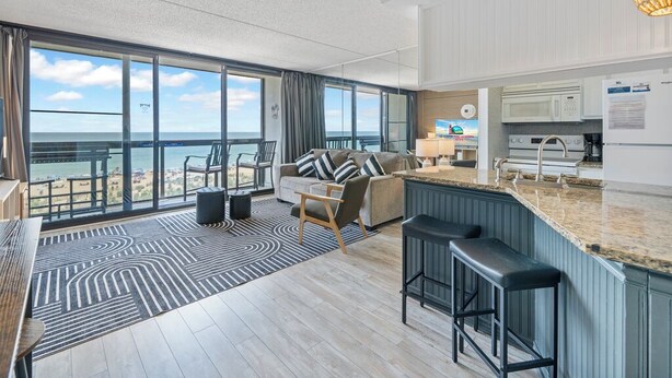 A modern hotel room with a large balcony, a sofa, a dining area, and a kitchen. - Family Friendly condo w/ Pools by Seaside Vacations & Sales (Ocean City)