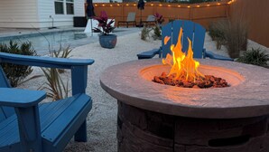 A backyard with a stone fire pit, blue Adirondack chairs, a pool, and string lights.