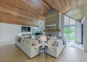 Een ruime woonkamer met een hoge nok, een open haard, een piano en een groot raam met uitzicht op de omgeving.