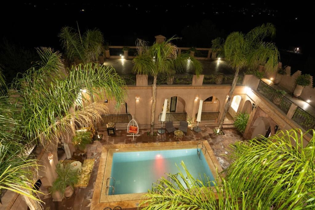 Zona de piscina en la azotea con una piscina rectangular, rodeada de palmeras y plantas en macetas. Hay zonas de estar al aire libre con mesas y sillas.