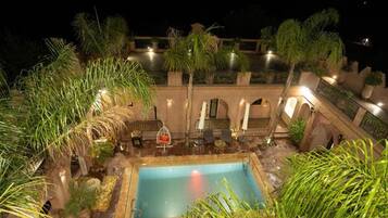 Zona de piscina en la azotea con una piscina rectangular, rodeada de palmeras y plantas en macetas. Hay zonas de estar al aire libre con mesas y sillas.