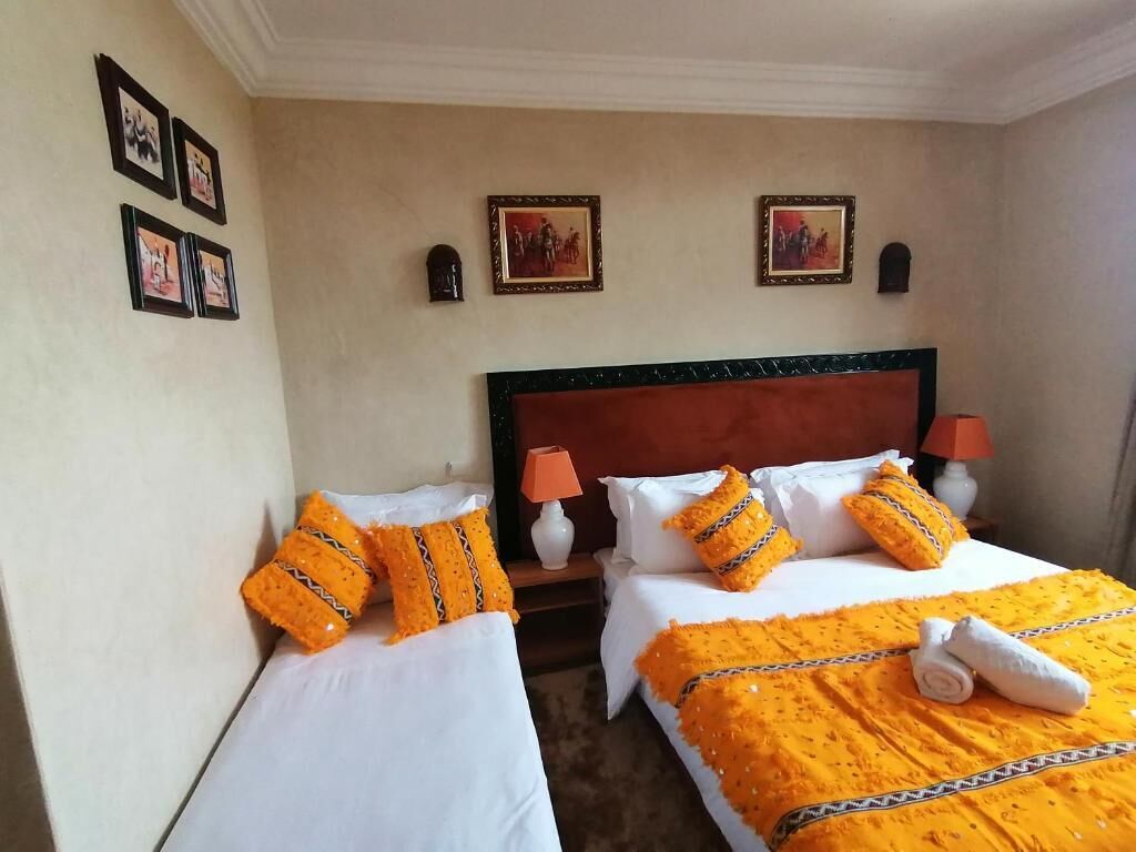 A hotel room with two beds, each with white linens and orange decorative pillows. There are framed pictures on the wall and a lamp on the nightstand.