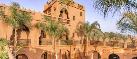A traditional building with multiple balconies, a swimming pool, and outdoor seating areas surrounded by palm trees.