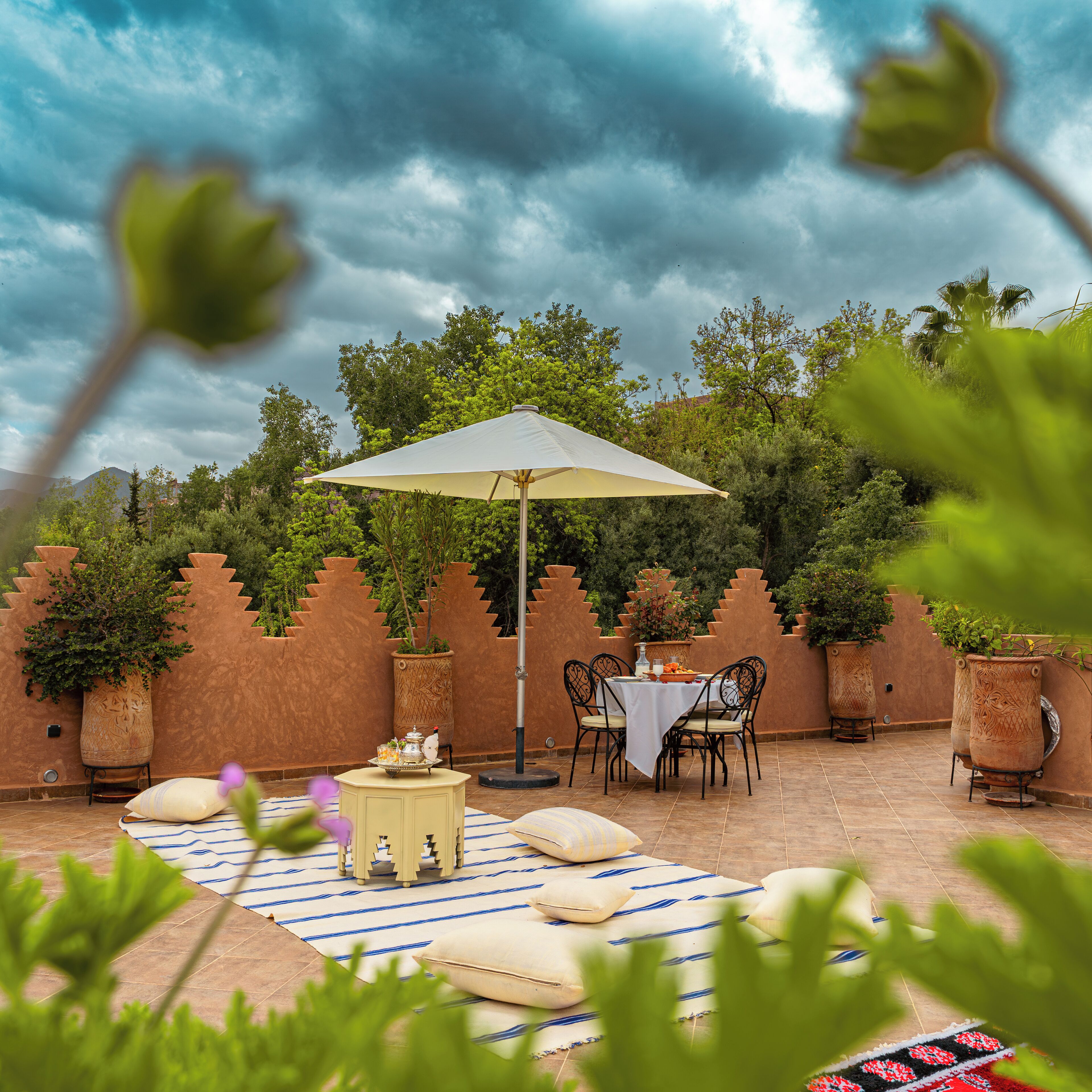 Un patio al aire libre con una mesa y sillas bajo una pérgola, rodeado de plantas en macetas y una pared decorativa.