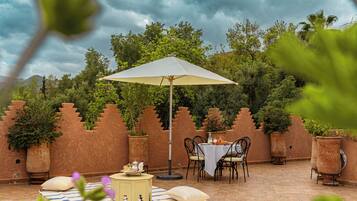 Un patio al aire libre con una mesa y sillas bajo una pérgola, rodeado de plantas en macetas y una pared decorativa.