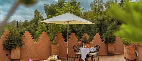 An outdoor patio with a table and chairs under a canopy, surrounded by potted plants and a decorative wall.