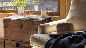 A cozy room with a large window, a wooden chest, a cushioned chair, and a small table with a vase of flowers, glasses, and a cup.