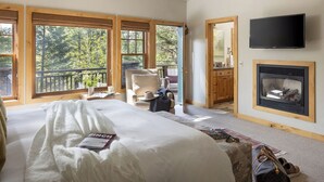 A bedroom with a large bed, a fireplace, a TV, and a balcony with a view of trees.