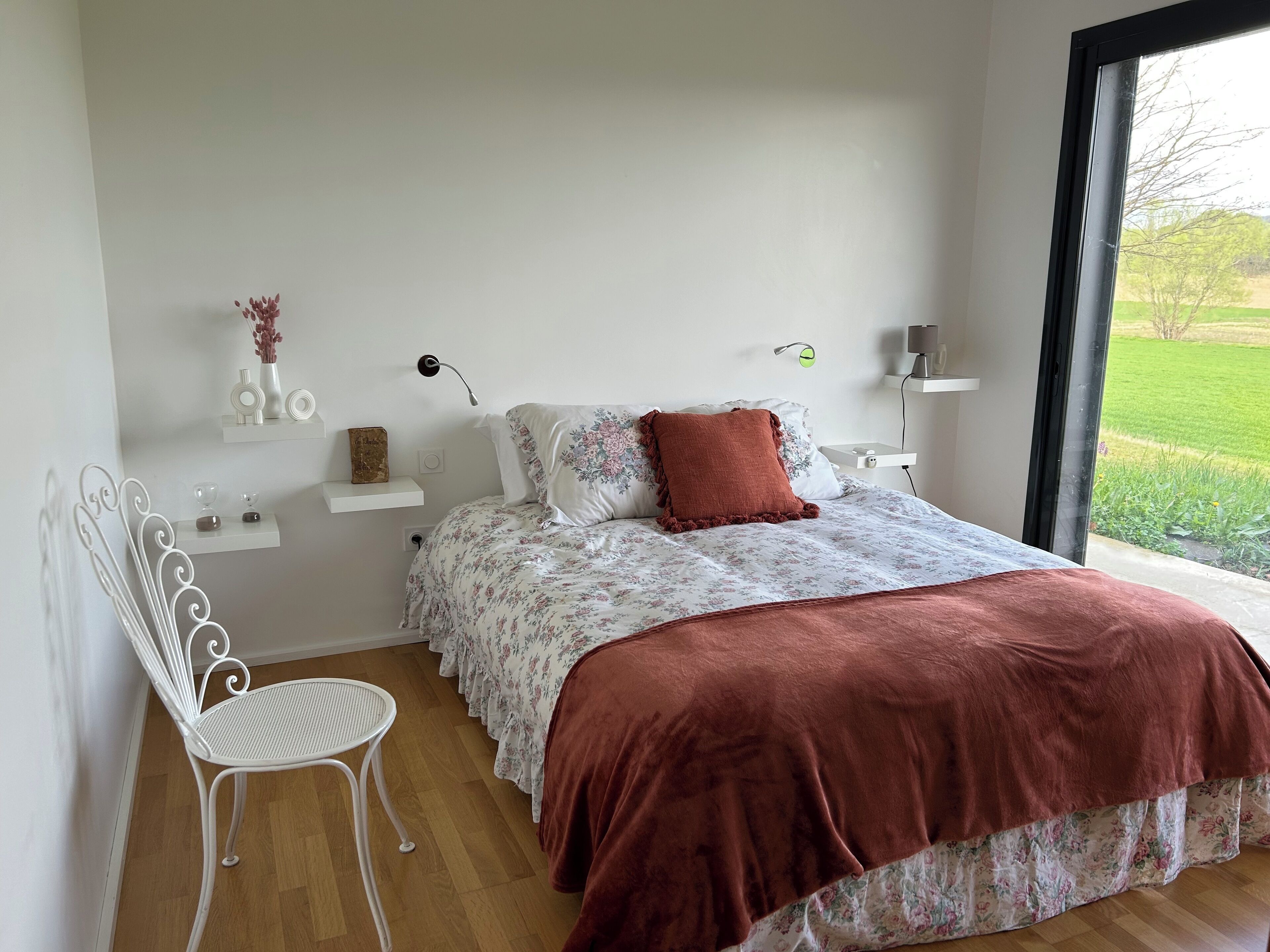 A bedroom with a bed, a chair, a bedside table, and a window with a view of greenery.