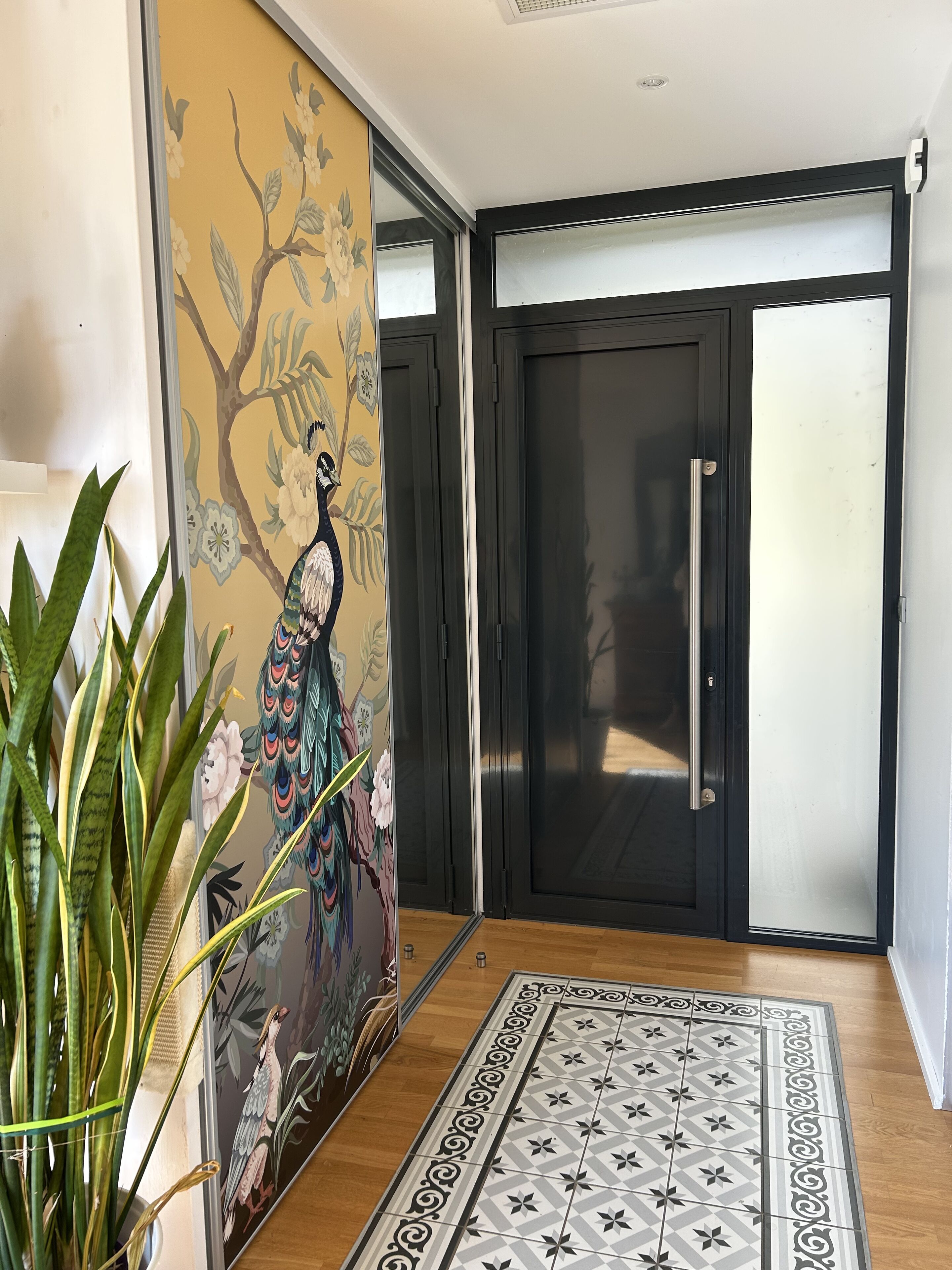 A room with a sliding door, a decorative wall with a peacock mural, a patterned rug, and a green plant.