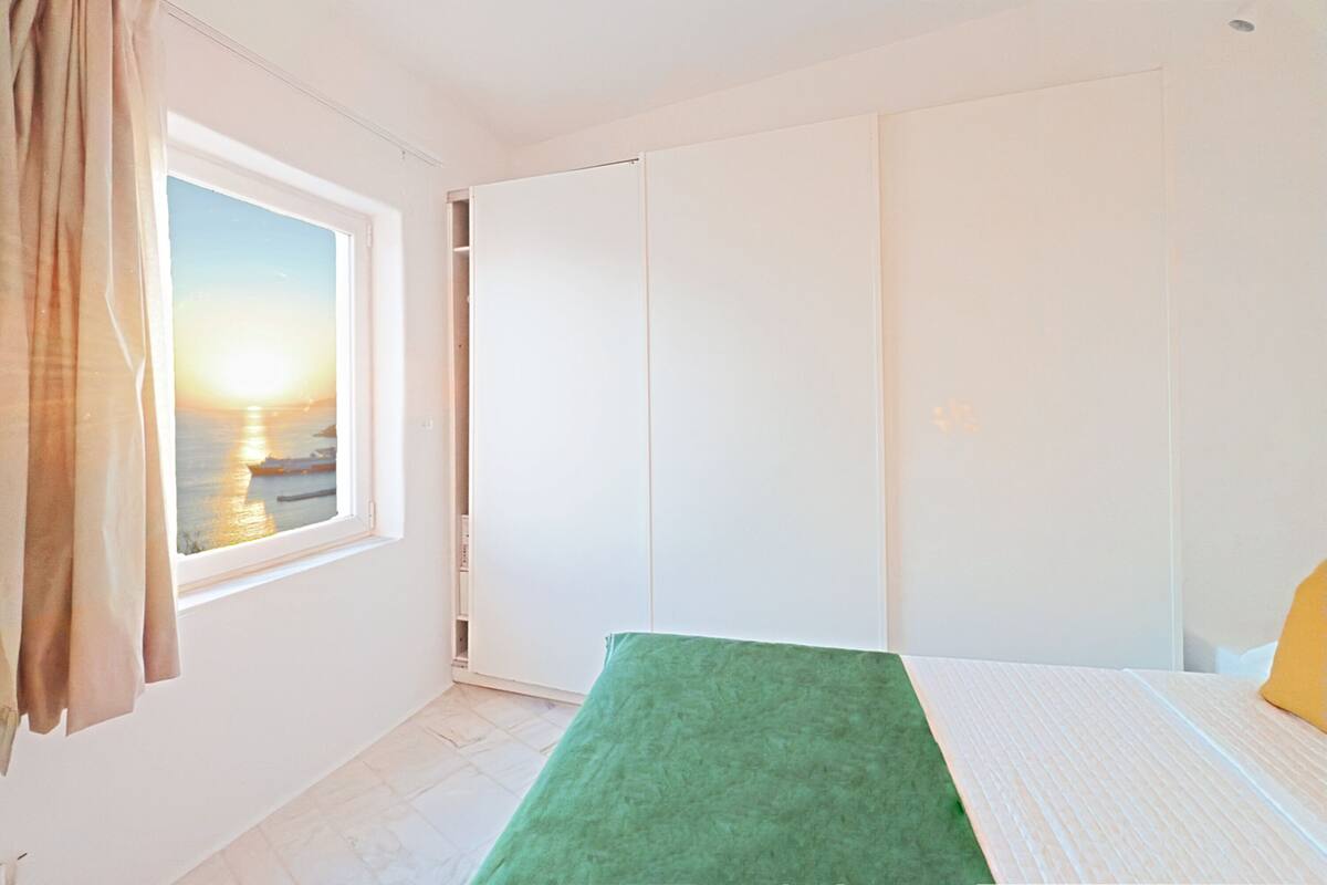 Une chambre à coucher avec un lit, une fenêtre avec des rideaux, une armoire et une vue sur le coucher de soleil sur l’eau.