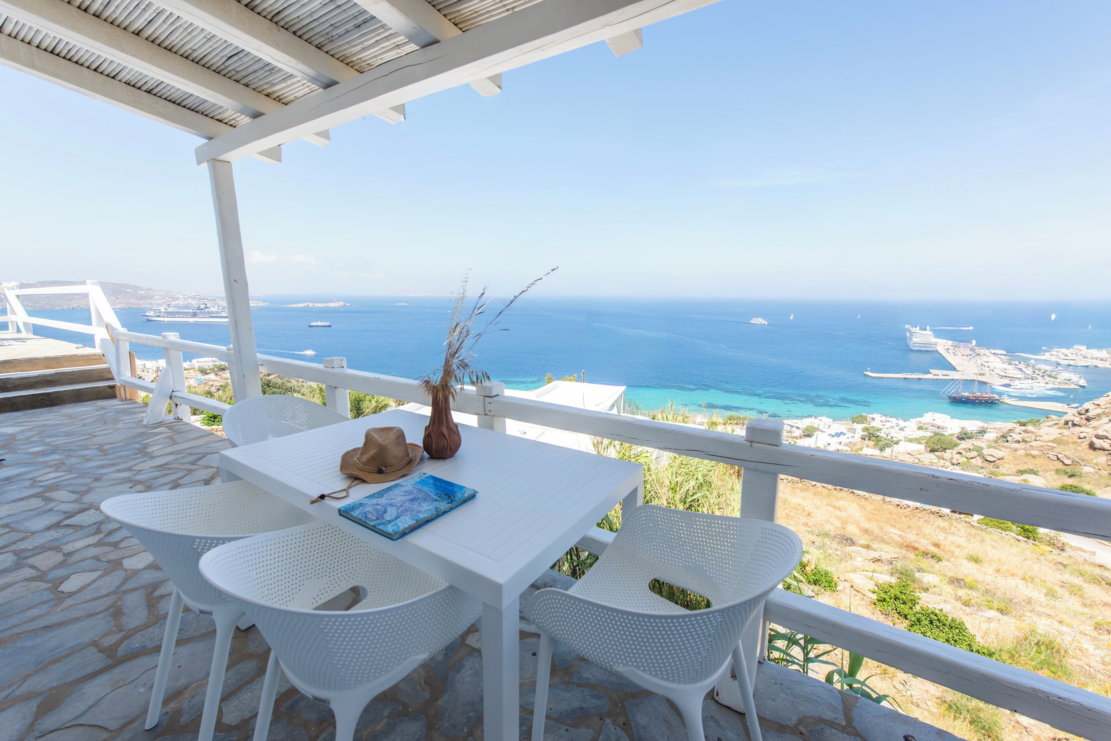 A terrace with a white table and chairs, overlooking a coastal view with boats and a beach.