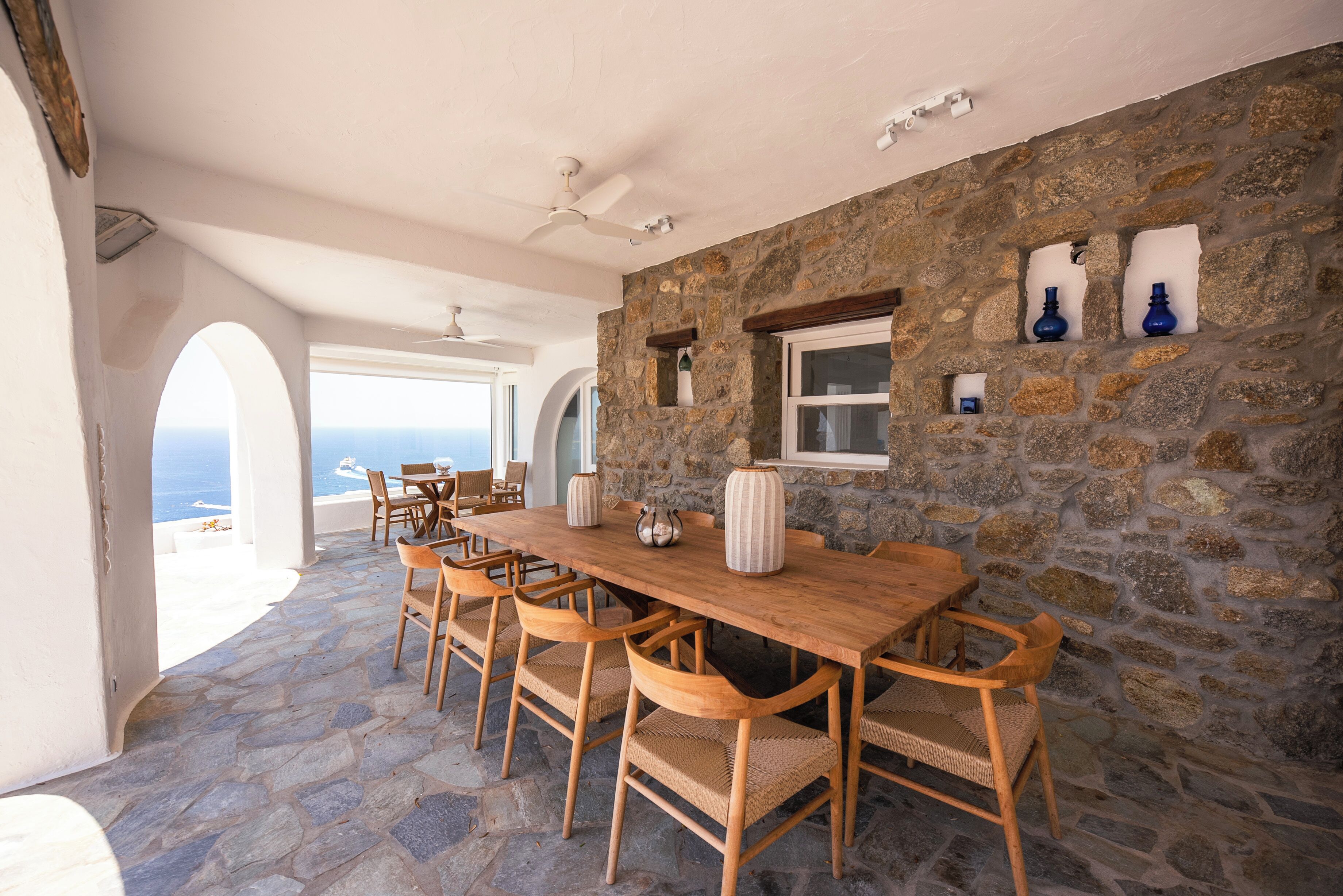A stone-walled dining area with a wooden table and chairs, a ceiling fan, and a view of the sea.