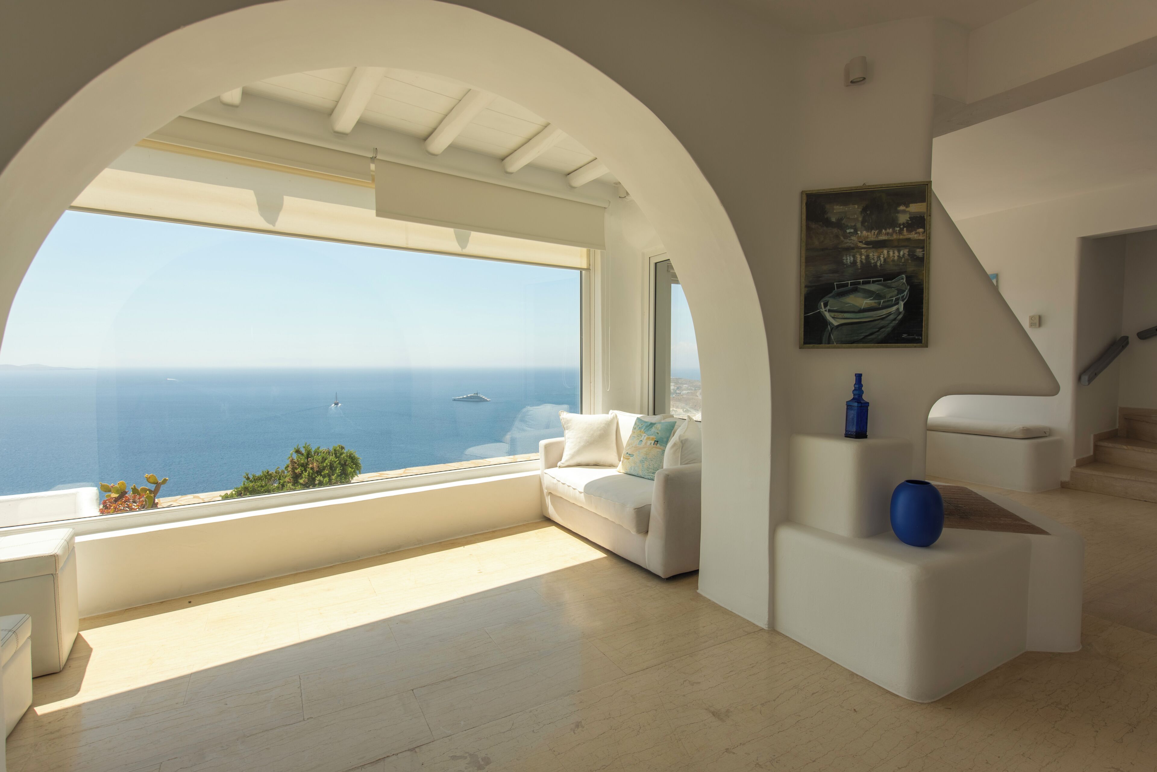 A modern interior with a large window offering a view of the sea, a white sofa, and a staircase.