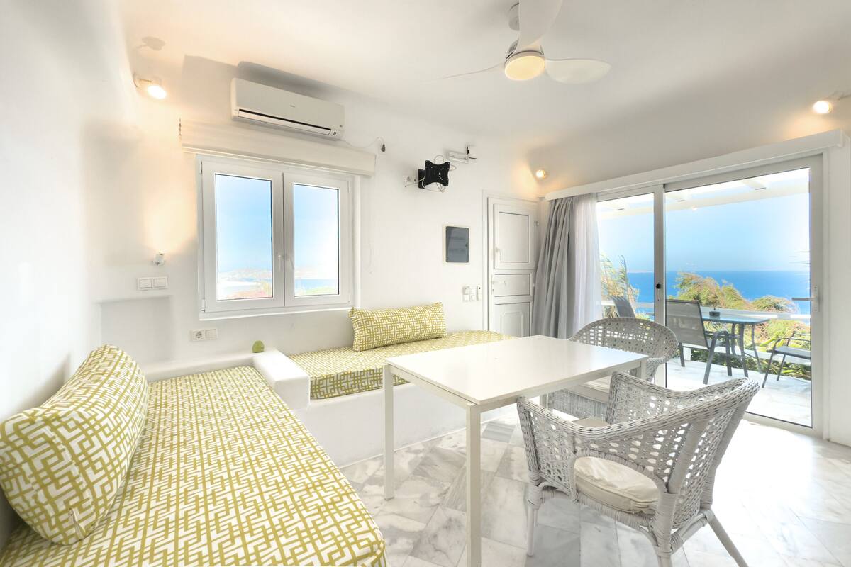 Une pièce lumineuse avec un canapé blanc, une table à manger, des chaises en osier et une vue sur la mer grâce à de grandes fenêtres.
