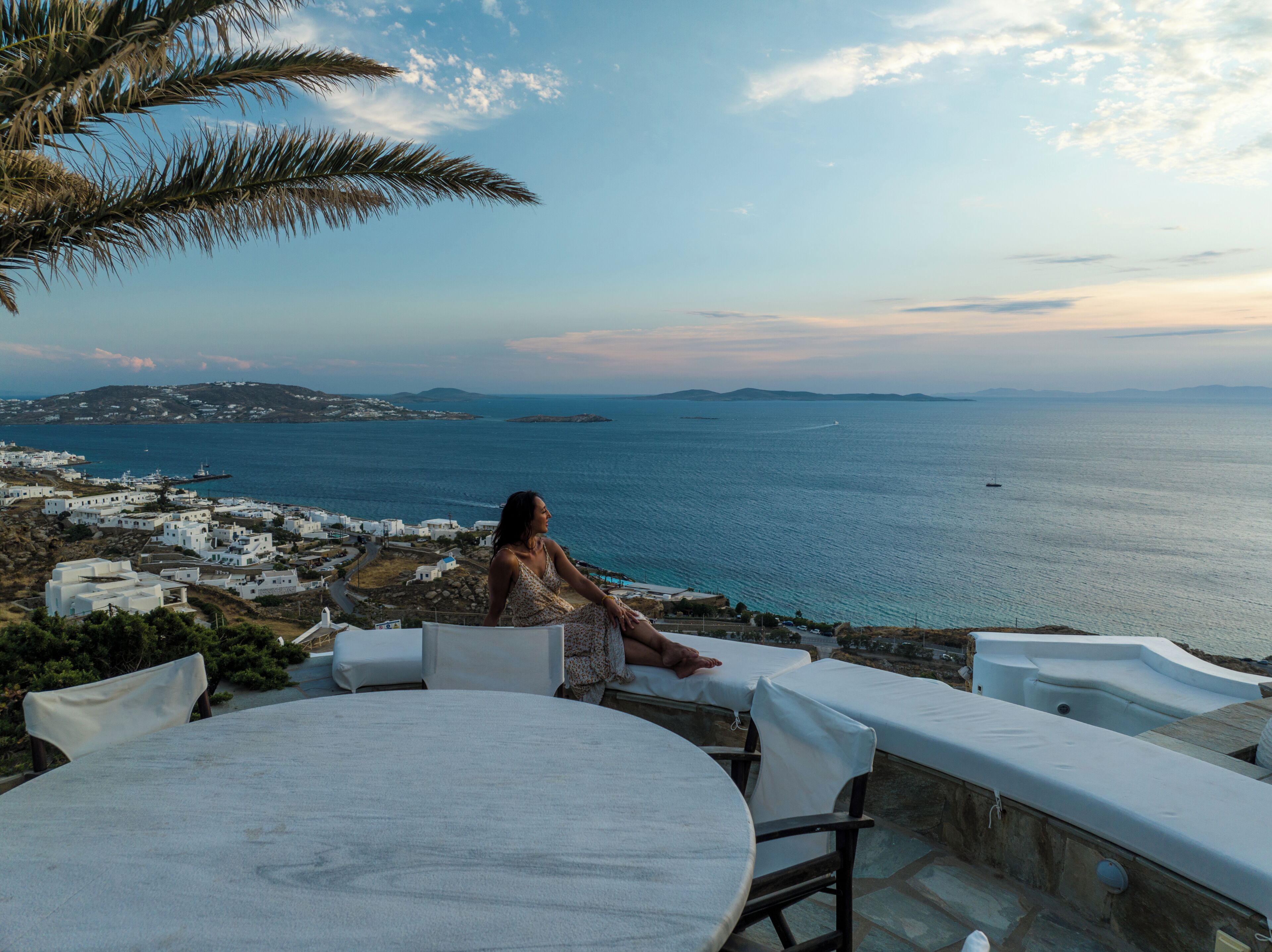 Vista costeira com uma mulher sentada à mesa, apreciando o mar, edifícios brancos ao fundo e céu claro.