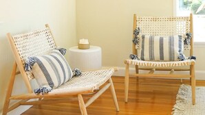 A room with two wicker chairs, a small round table, and a window with a view of greenery. - The Captain's Cottage on Shoreline Drive (Santa Barbara)