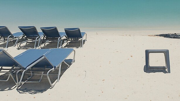 Une plage bordée de rangées de transats et comportant une seule chaise vide, un ciel bleu dégagé et une mer calme.