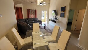 Un salon avec un canapé en cuir noir, une table basse en verre et un téléviseur posé sur un meuble. On y trouve également un coin repas avec une table et des chaises en verre.