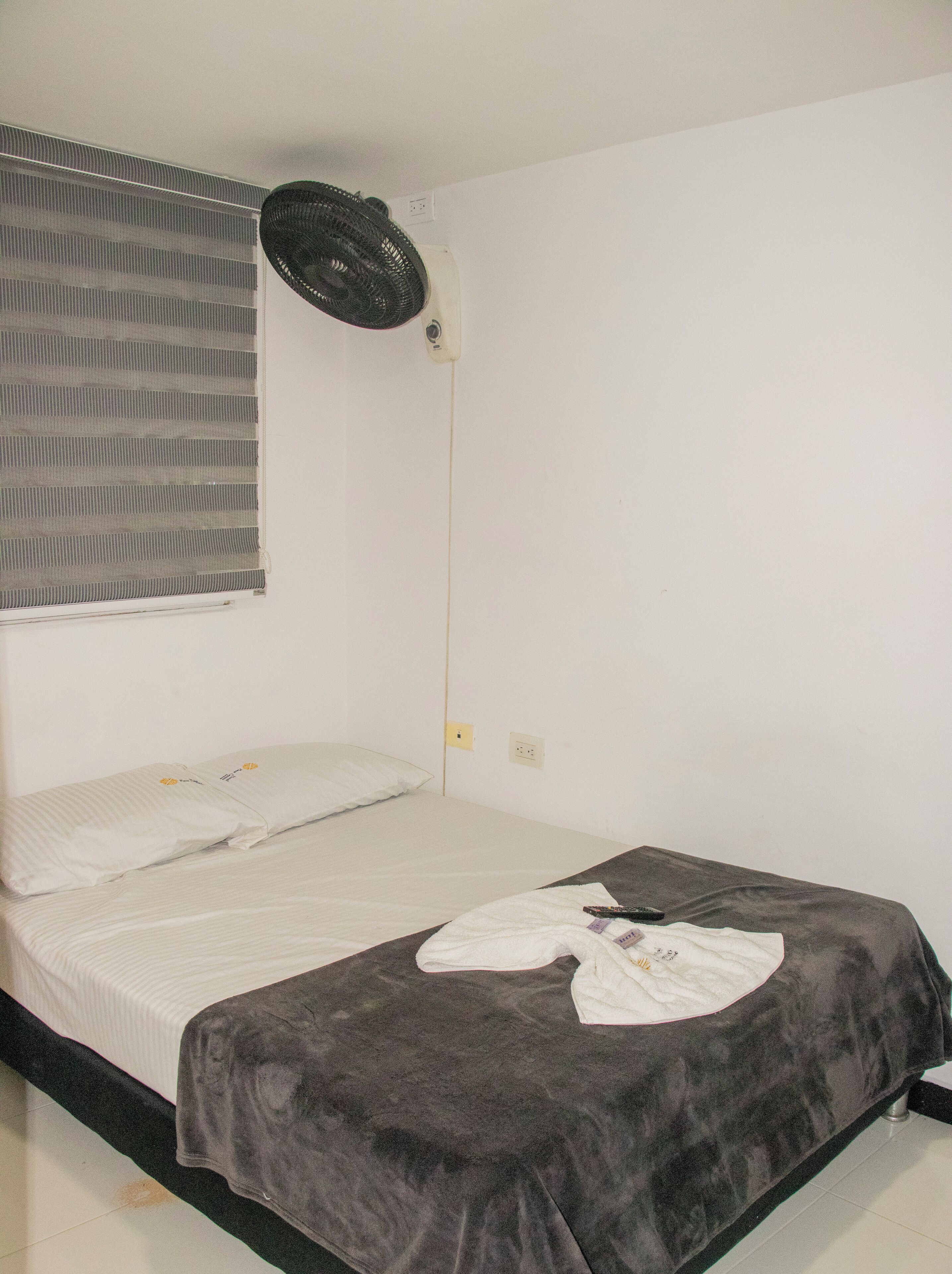 Ein Einzelbett mit grauer Decke und Kissen, ein Standventilator, ein Fenster mit Rollo sowie eine Steckdose an der Wand.
