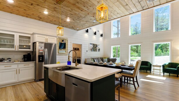 A modern kitchen with a central island, stainless steel appliances, and a dining area with green upholstered chairs. - NEW Listing! Elevated forest views at Pines Peak! (Broken Bow)