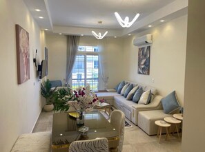 A modern living room with a sofa, a dining table with chairs, a coffee table with a vase of flowers, and a large window with curtains. - Deluxe Apartment in the North Coast  (New Alamein)