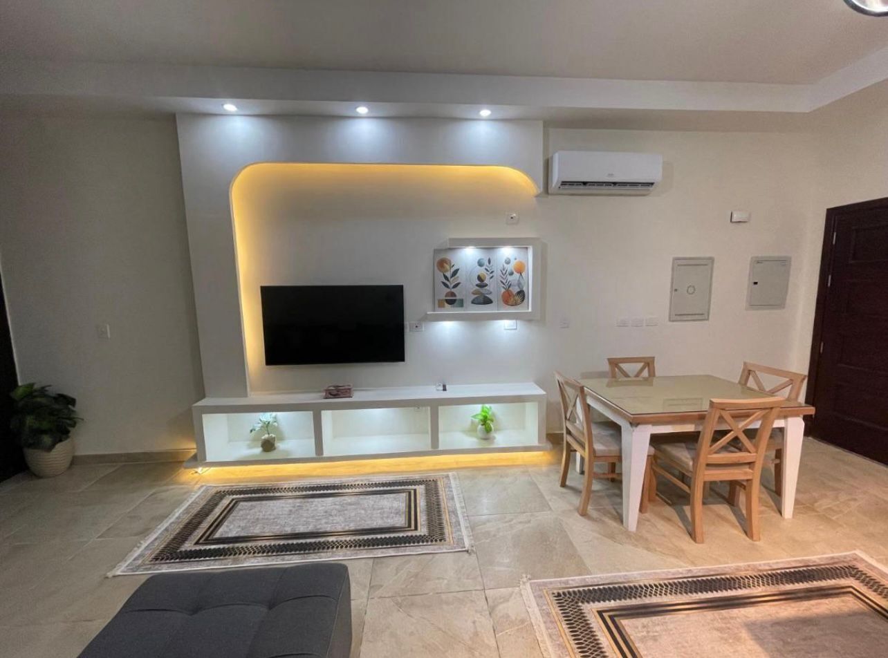 Una sala moderna con un televisor de pantalla plana, una mesa de comedor con sillas, un sofá y una estantería decorativa con plantas.