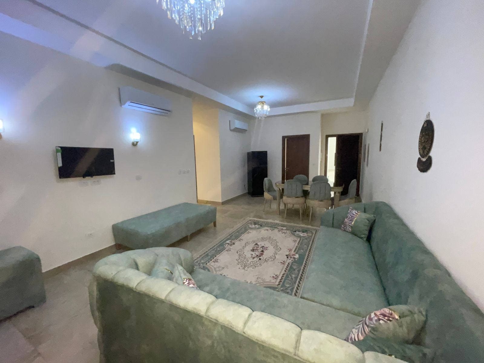 Ein Wohnzimmer mit einem grünen Sofa, dazu passendem Hocker, einem Esstisch mit Stühlen, einer Kristallleuchter und einem an der Wand montierten Flachbildfernseher.