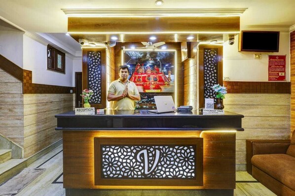 A reception desk with a laptop, a sign indicating "CHECKOUT TIME," and a decorative wall with a large letter 'V'. - Hotel Vinayak -Banganga Road Katra  (Reasi)