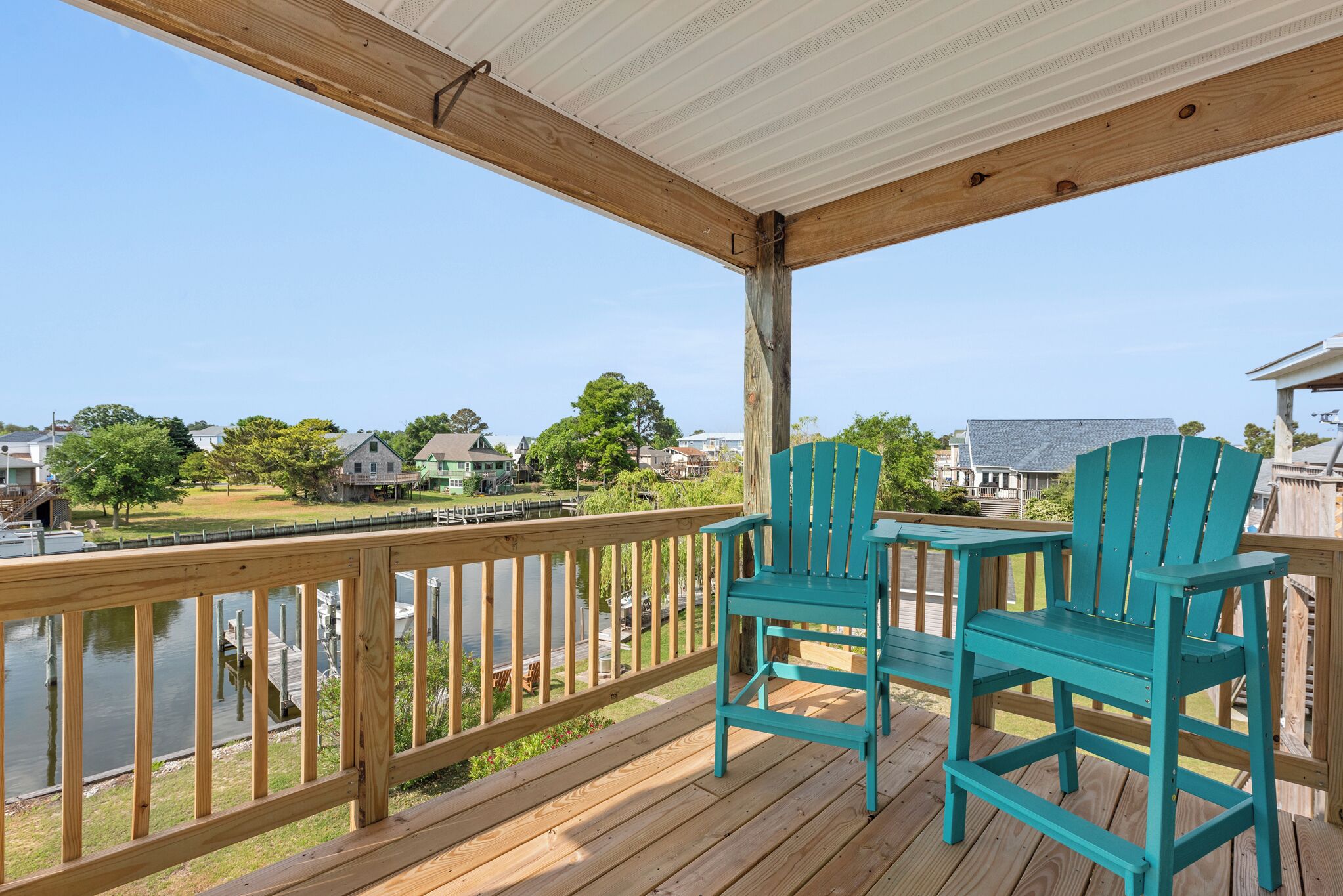 A wooden deck with two turquoise Adirondack chairs overlooking a body of water and houses.