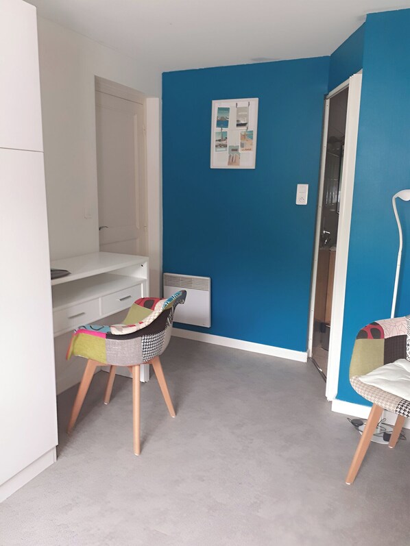 A small, bright room with a blue accent wall, a white desk, two chairs, and a radiator. - Quiet T1 in St Marc sur Mer, 5 min walk to the beach, terrace and large bed! (Saint-Nazaire)