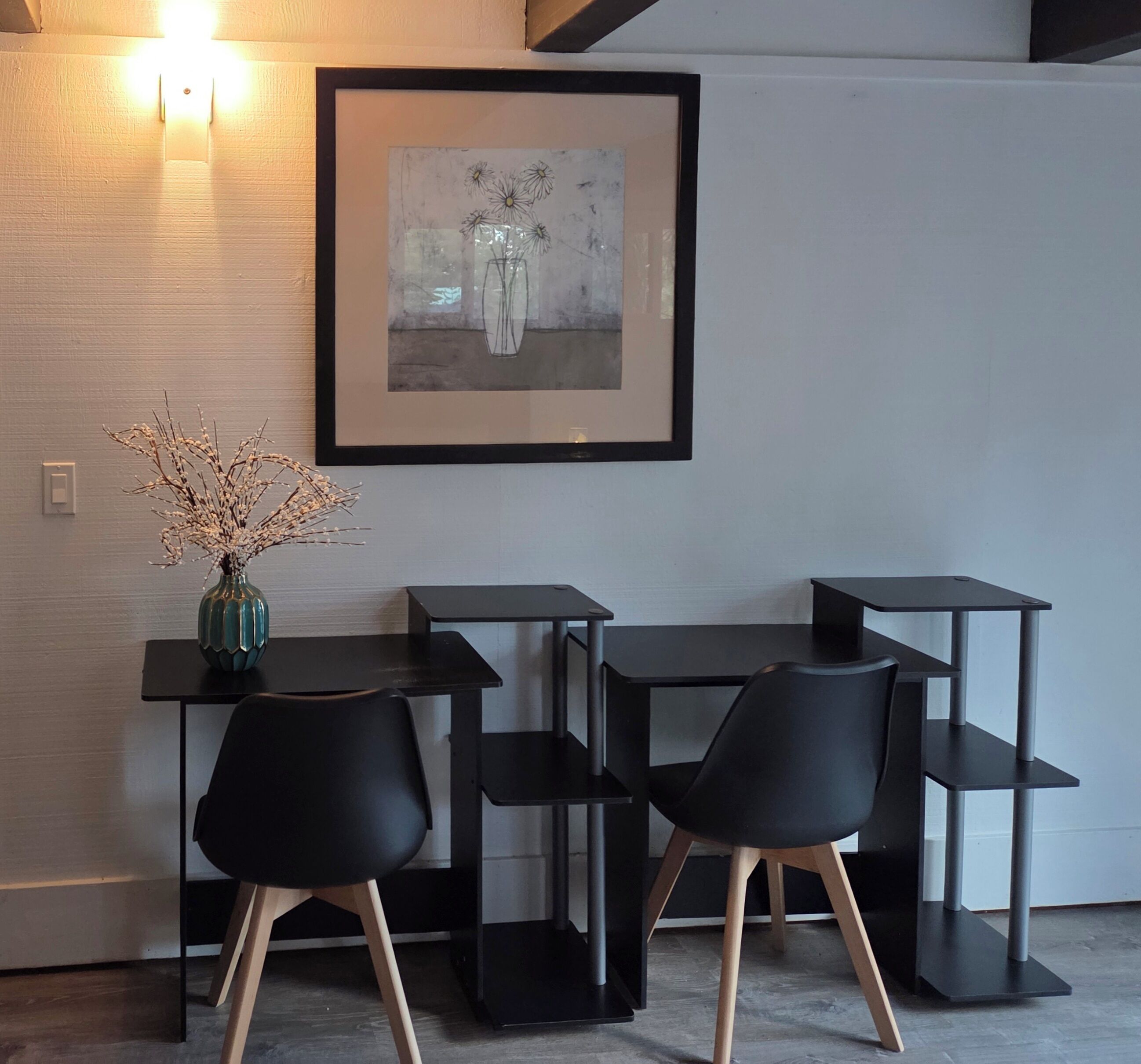 A modern room with a desk and two chairs, a framed artwork on the wall, and a vase with branches on the desk.