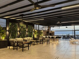 Un área de estar moderna con techo alto, vigas a la vista y un amplio ventanal con vistas al mar. Hay varios sofás y sillones dispuestos para sentarse, además de un pequeño bar con mostrador de piedra.