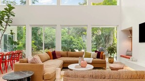 A spacious living room with a large sectional sofa, a round coffee table, and a woven pouf. There are large windows offering a view of greenery outside. - Lake Austin Luxury/200ft Private Waterfront on 2 Acres/2 Docks/Sleeps 4 (Austin)