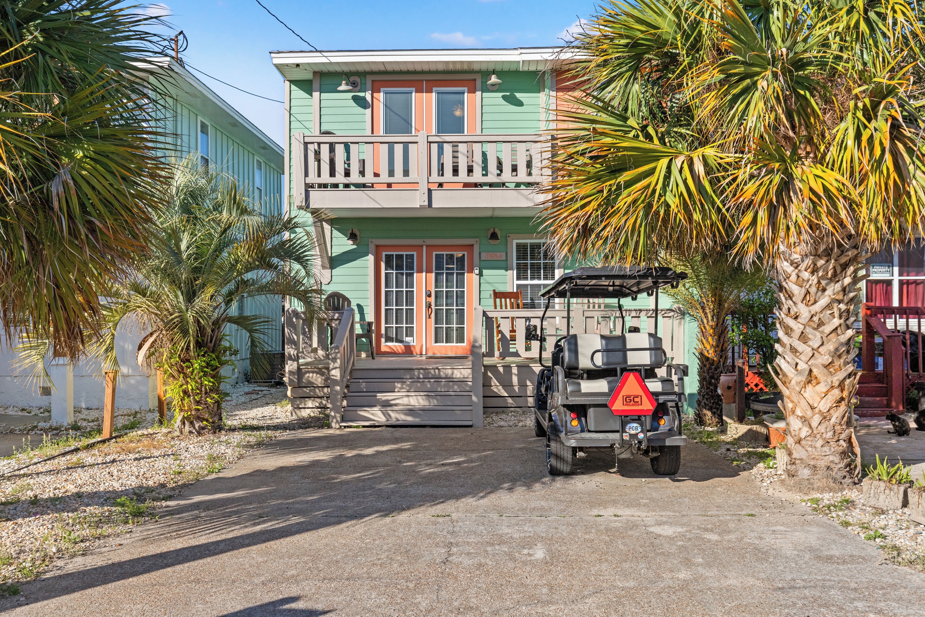 A two-story building with a balcony, a vehicle parked in front, and palm trees.