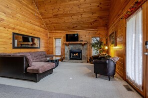 L’intérieur d’un chalet confortable, avec des murs en bois, une cheminée en pierre, un téléviseur fixé au-dessus et un coin salon accueillant, comprenant un canapé et des fauteuils.