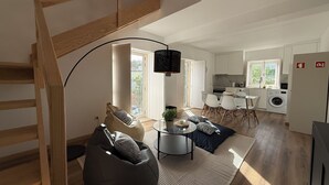Ett modernt vardagsrum med en trätrappa, en svart sittpuff, ett runt soffbord och en matplats med ett glasbord och stolar.