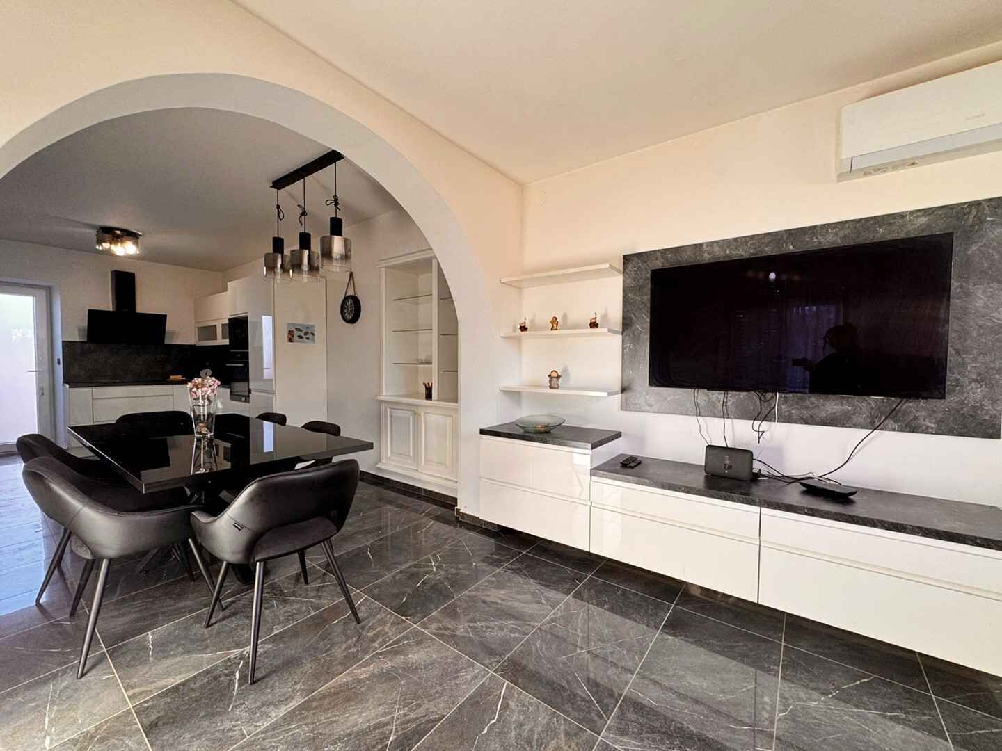 A modern kitchen with a dining area, featuring a large TV, white cabinetry, and a marble floor.