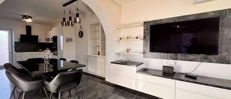 A modern kitchen with a dining area, featuring a large TV, white cabinetry, and a marble floor.