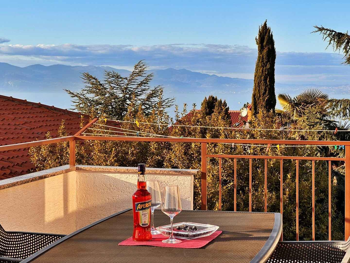 Un balcón con una mesa, una botella de Aperol y dos vasos, con vistas a un paisaje montañoso.