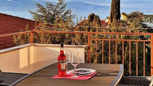 Uma varanda com uma mesa, uma garrafa de Aperol e dois copos, com vista para uma paisagem montanhosa.