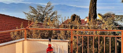 A balcony with a table, a bottle of Aperol, and two glasses, overlooking a mountainous landscape.
